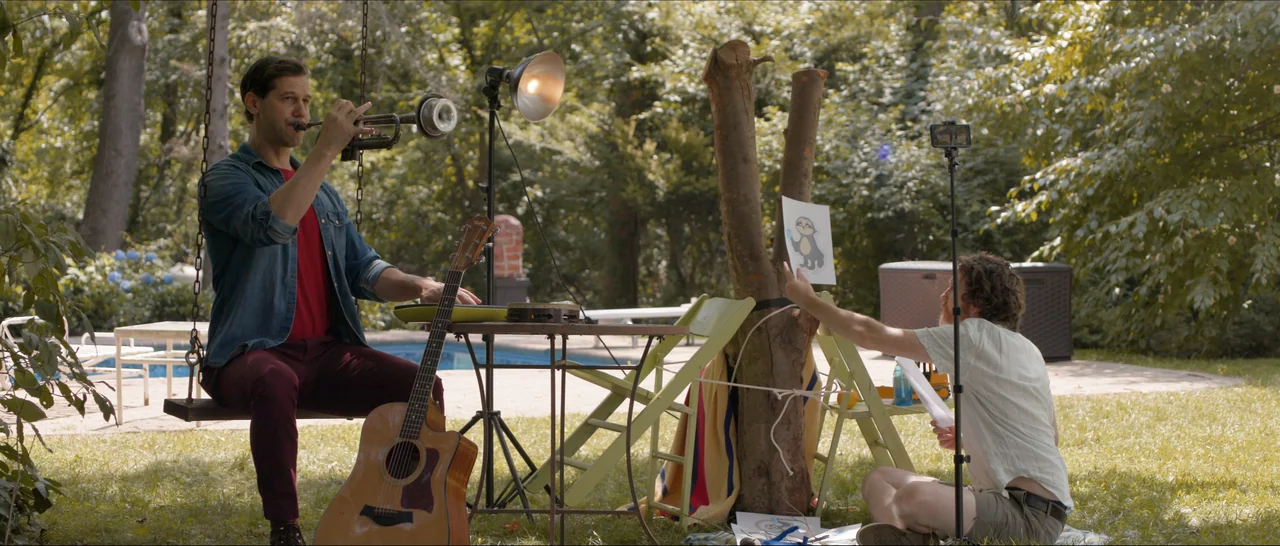 Songs For A Sloth (2021) movie still: wide — A grassy area with a swimming pool in the background features trees and lawn furniture. One man plays…