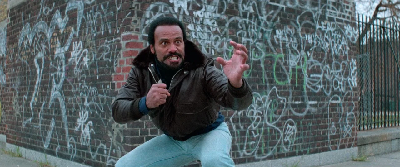Vigilante (1982) movie still: medium — A corner of a red brick building covered in dense white graffiti tags. A man crouches in a defensive martial…