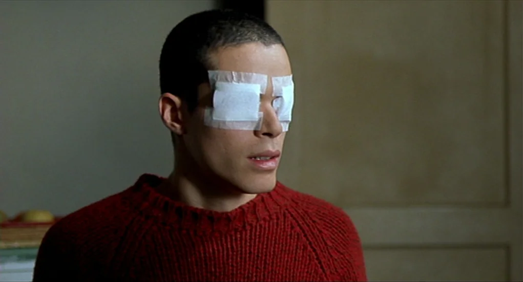 Tiresia (2003) movie still: closeup — A man stands in an indoor space with plain walls and a simple door frame. He wears a bandage covering both of…
