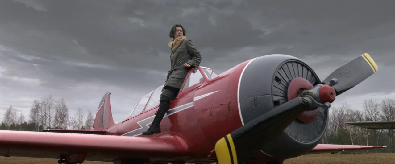 Resident Evil: Afterlife (2010) movie still: fullbody — An overcast sky hangs over a barren, grassy field with a single red aircraft. A woman stands…