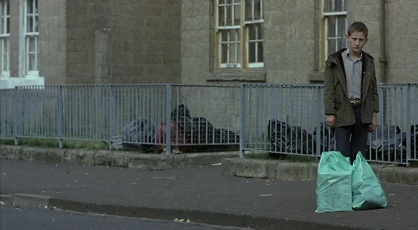 Ratcatcher (1999) movie still: fullbody — A sidewalk borders a stone building and a metal fence filled with trash bags. A young man stands on the…