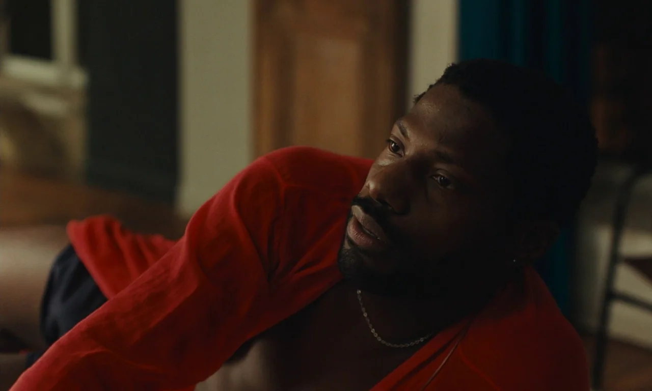 Passages (2023) movie still: closeup — A dimly lit room with a soft-focus background of muted walls and furniture. A man in a red shirt reclines,…