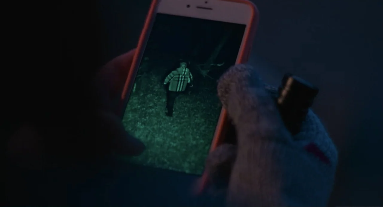 Turtles All the Way Down (2024) movie still: closeup — A close-up view of a person wearing knit gloves holding a modern smartphone outdoors. The…