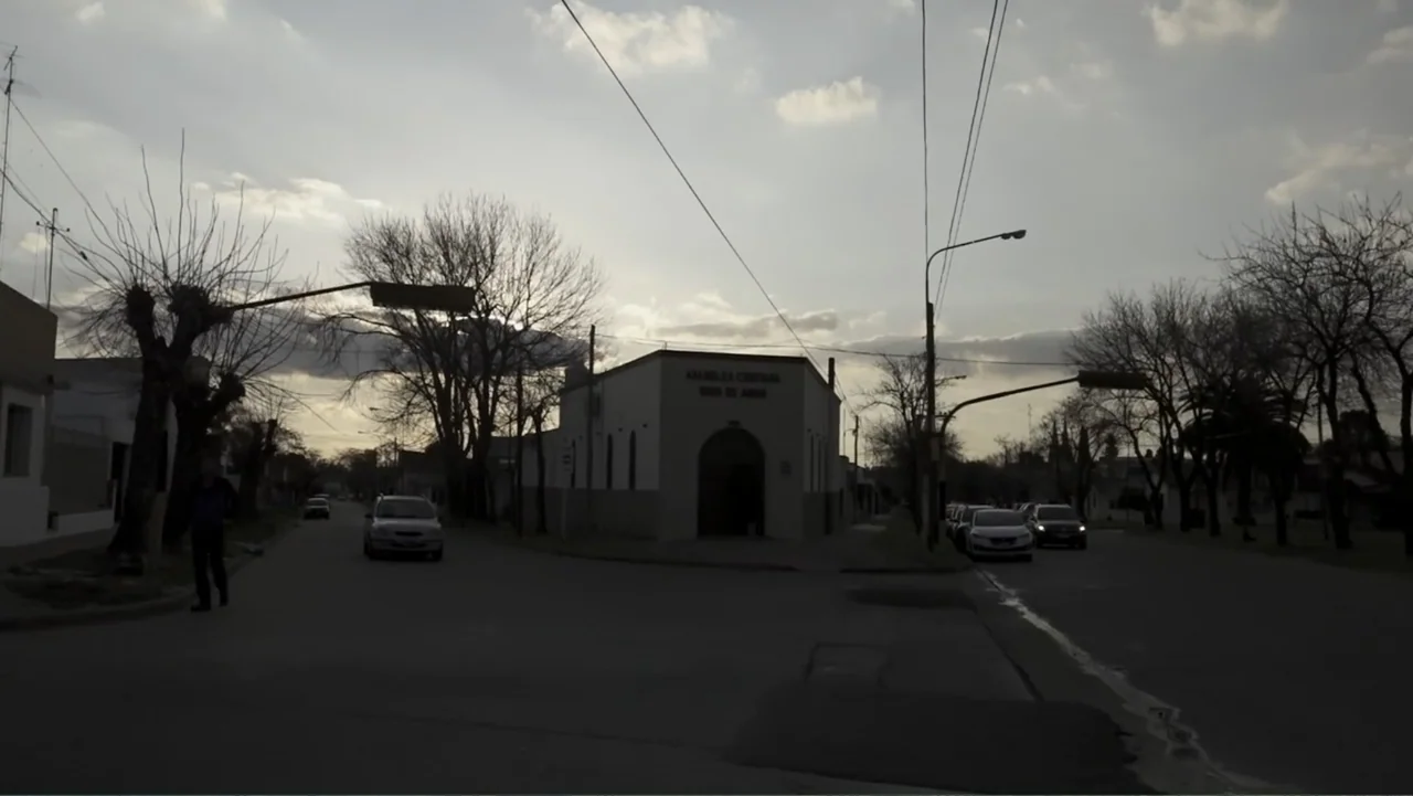 Saturdays Disorders (2022) movie still: establishing — An intersection in a small town features a white church at the center. Several cars drive on…