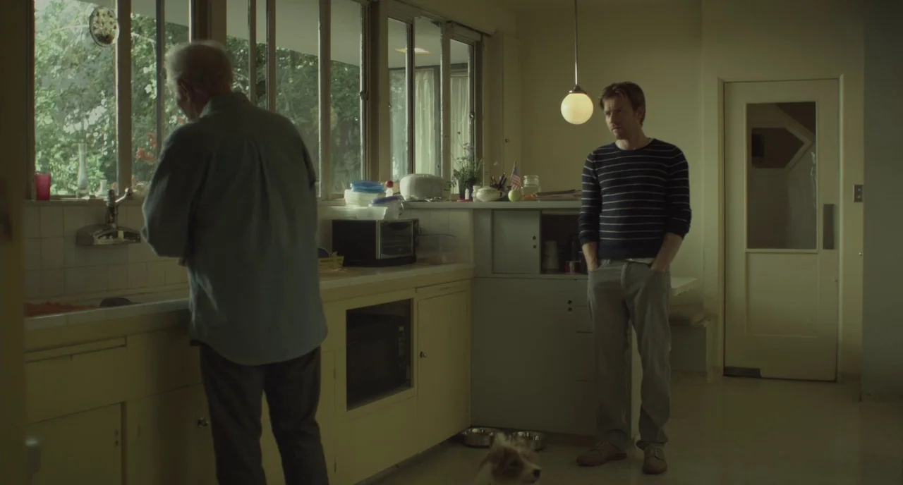 Beginners (2010) movie still: medium — A residential room features yellow cabinetry, a long row of windows, and a tiled floor. One man washes dishes…