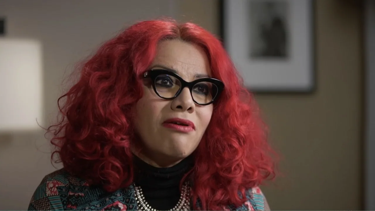 Escape From Saudi (2019) movie still: closeup — A person with bright red hair sits in a sparsely furnished indoor room. The subject looks toward the…