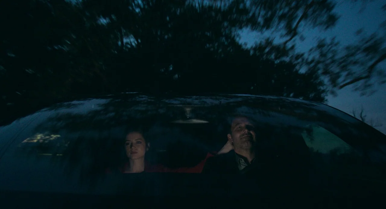 Deep Water (2022) movie still: medium — A view through a car windshield of two people driving at night. Motion blur of dark trees lines the top of…