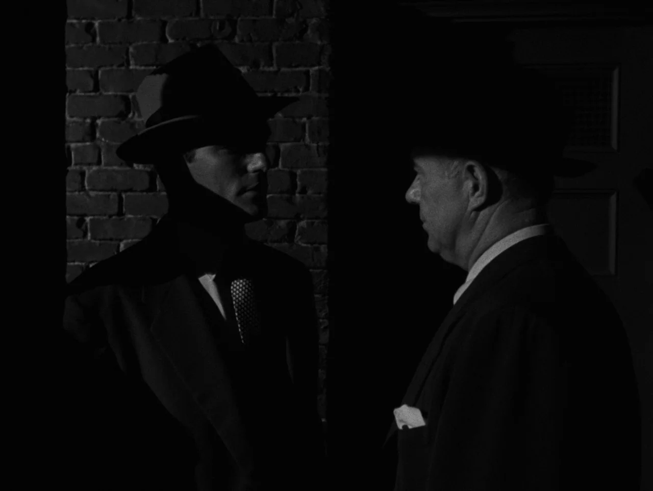 Pickup on South Street (1953) movie still: medium — Two men stand together at night against a dark brick wall surface. They face each other while…