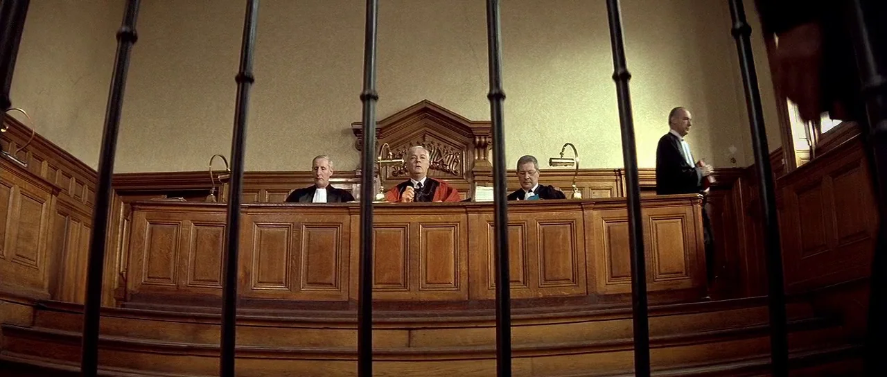 Nikita (1990) movie still: wide — A wood-paneled courtroom features three judges seated behind a high bench. Vertical metal bars frame the…