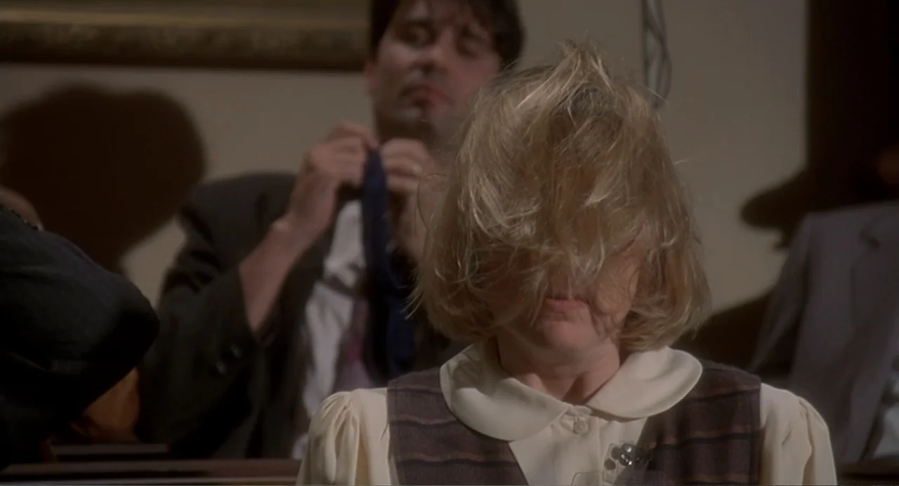 Fatal Instinct (1993) movie still: medium — A courtroom interior with rows of wooden benches in a dimly lit space. A woman sits in the foreground…