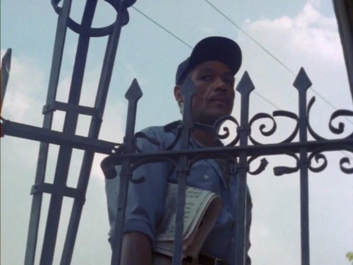 Tenement (1985) movie still: medium — A man stands outdoors behind a decorative black wrought iron fence under an overcast sky. He holds a folded…