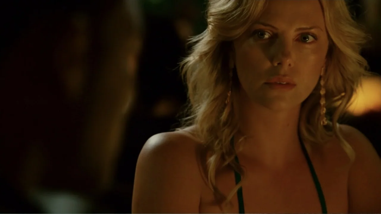Hancock (2008) movie still: closeup — A dimly lit indoor space with an out-of-focus foreground figure. A woman with blonde hair looks toward an…