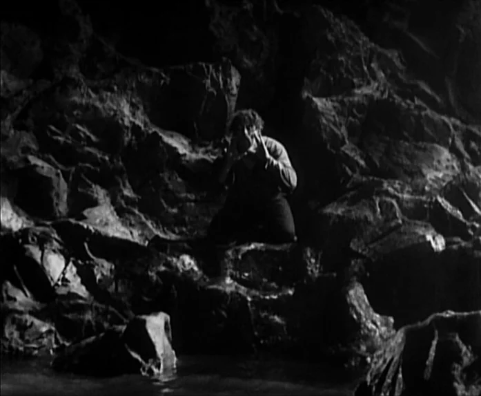 Sunrise (1927) movie still: wide — A dark, jagged cave interior features uneven rock surfaces and a small pool. A man kneels on a rock outcropping…