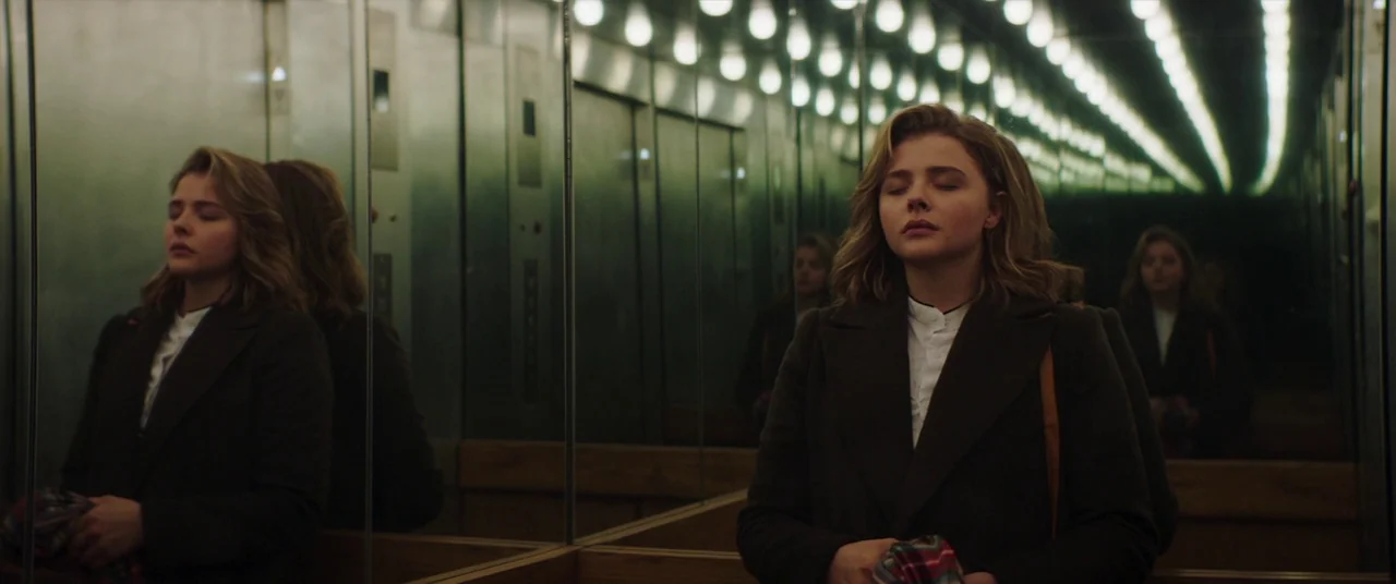 Greta movie still: medium — A person stands inside a mirrored car with panels of vertical steel and wood. The subject keeps their eyes closed while…