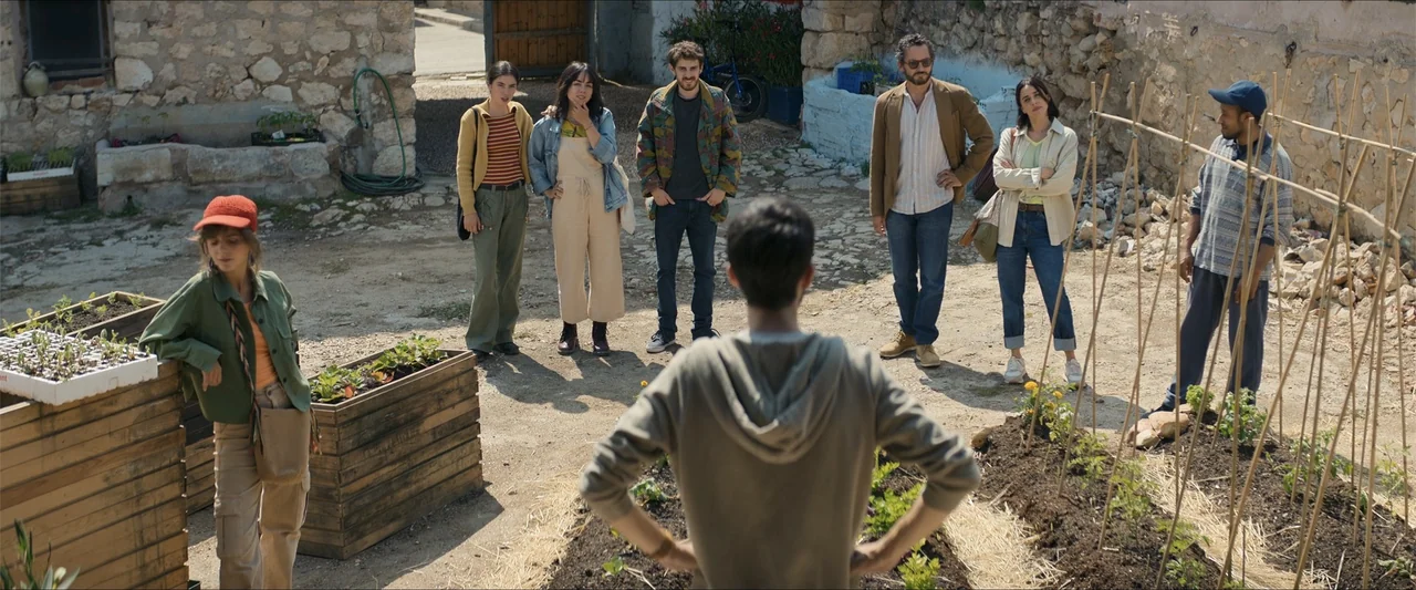 A Hipster in Rural Spain (2024) movie still: fullbody — An outdoor courtyard contains raised wooden planting beds and stone masonry walls. A group…
