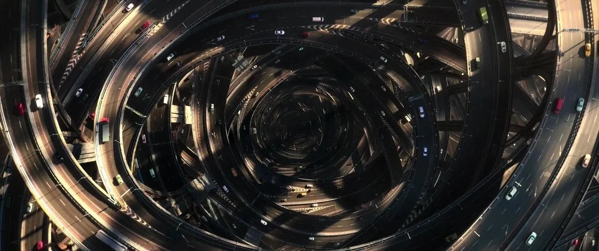 Three Thousand Years of Longing (2022) movie still: establishing — A complex multi-level highway interchange spirals into a dark, shadowed center…