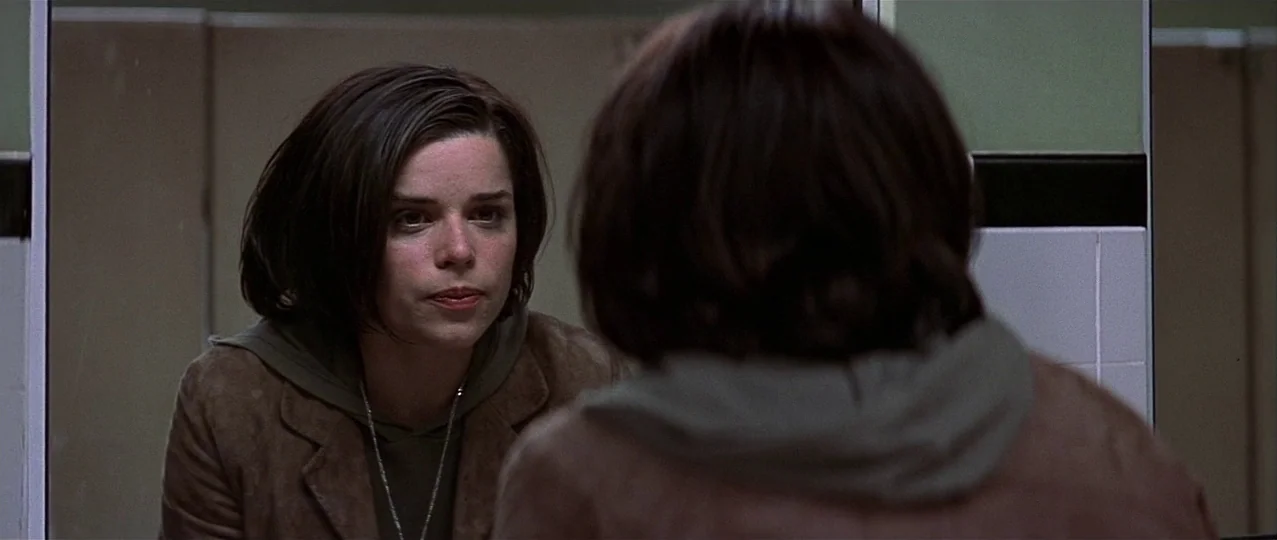 Scream 3 (2000) movie still: medium — A public restroom with light green tiled walls and dark trim dividers. A young woman views her reflection in…