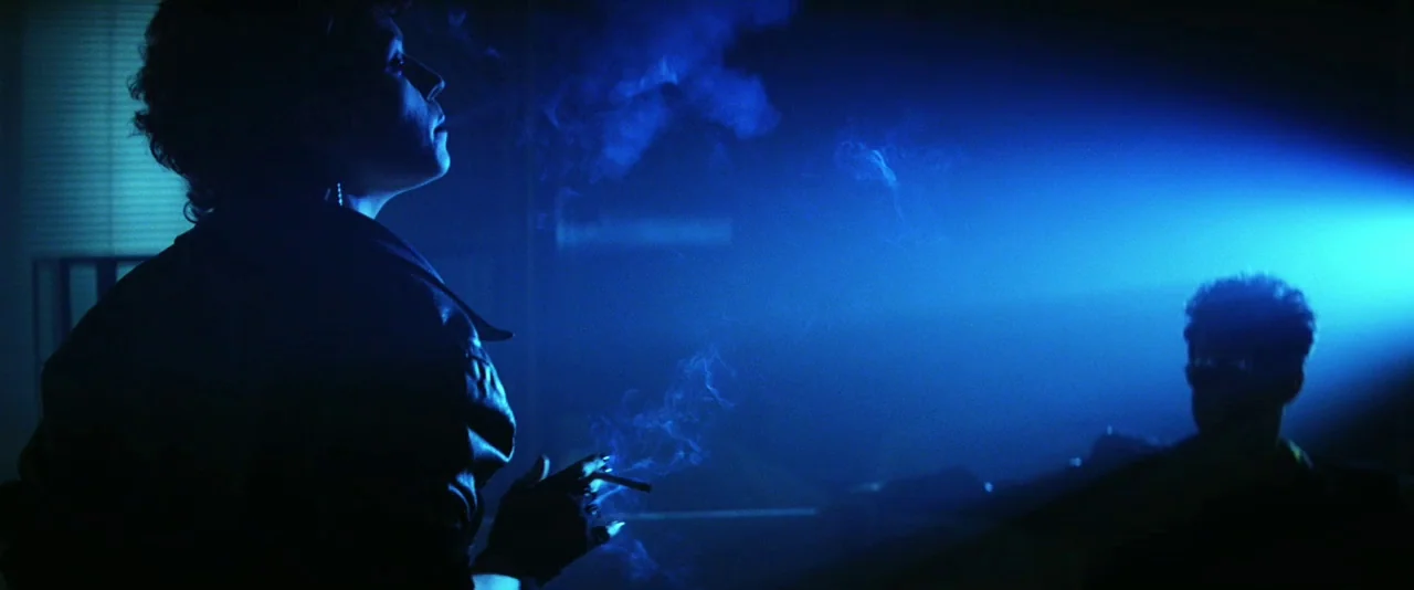The Hunger (1983) movie still: medium — A dimly lit room is bathed in intense blue light from a movie projector. A woman smokes in the foreground…