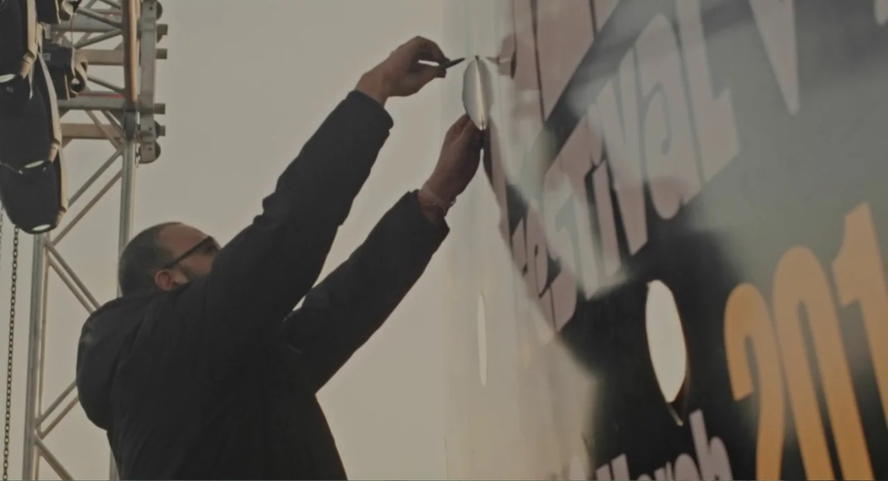 Cairo Jazzman (2017) movie still: medium — A man stands outdoors near a tall festival banner during late afternoon sun. He reaches upward with both…