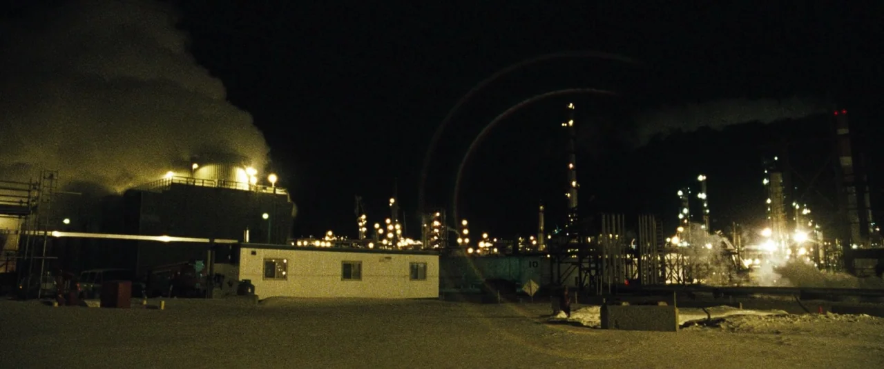 The Grey (2012) movie still: establishing — A nighttime view of an industrial refinery with heavy smoke rising into the air. Lit structures and…