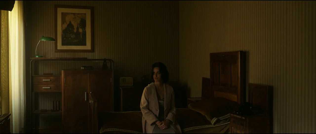 Ferrari (2023) movie still: medium — A dimly lit bedroom features wood furniture, striped wallpaper, and a framed picture on the wall. A woman…