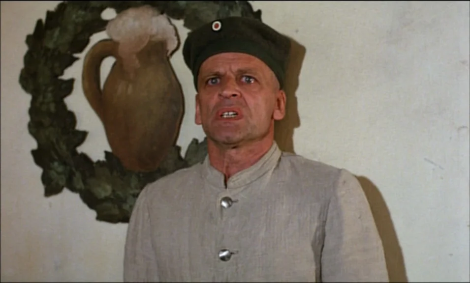 Woyzeck (1979) movie still: medium — A man stands indoors against a wall featuring a painted circular emblem of a beer stein. The subject faces the…
