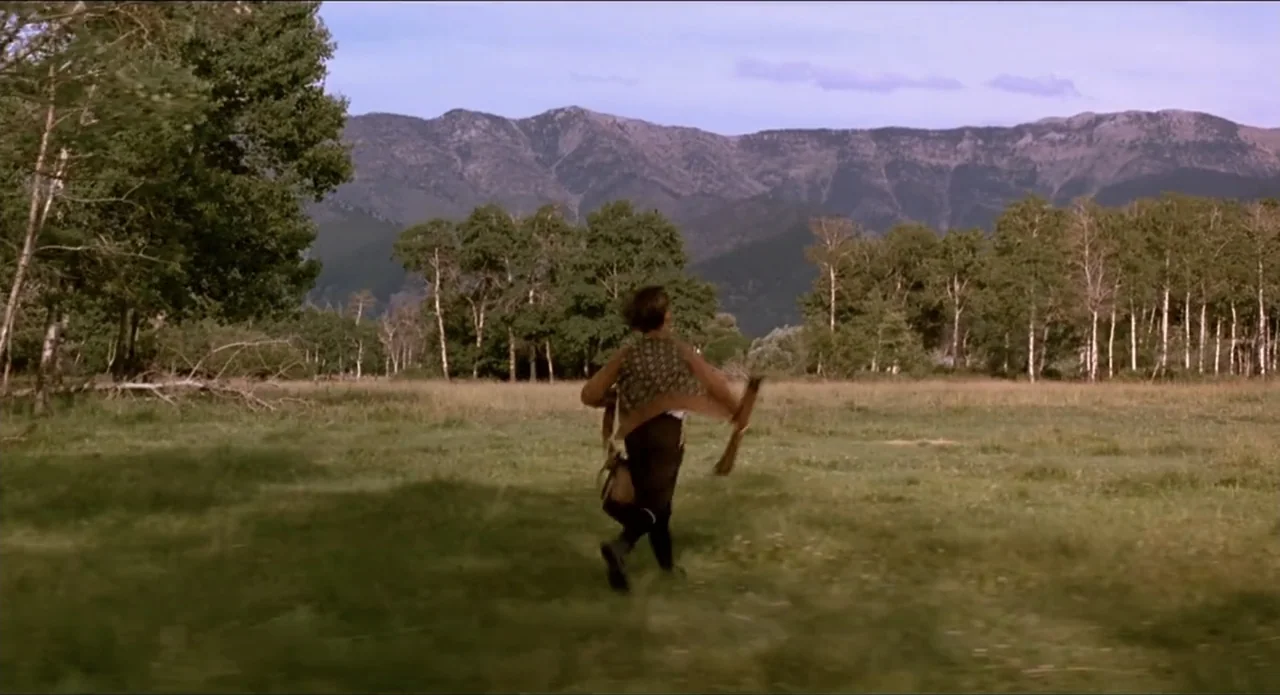 A River Runs Through It (1992) movie still: wide — A vast grassy meadow sits in front of a prominent mountain range. A young male runs away from the…