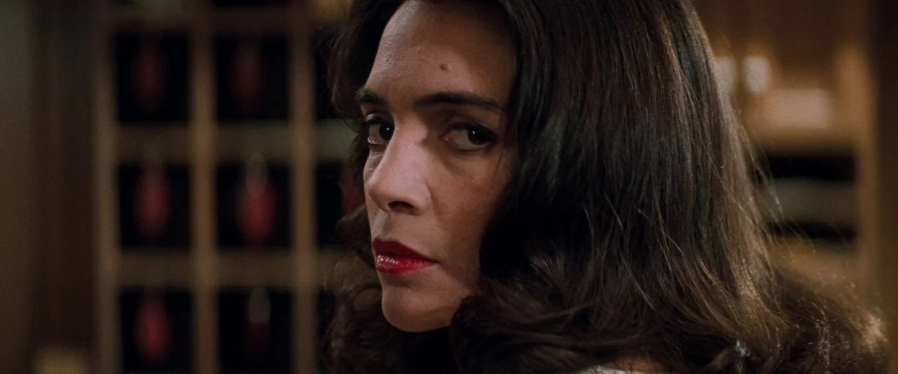 In The Mouth of Madness (1994) movie still: closeup — A dimly lit retail environment features shelves with red objects in the background. A woman…