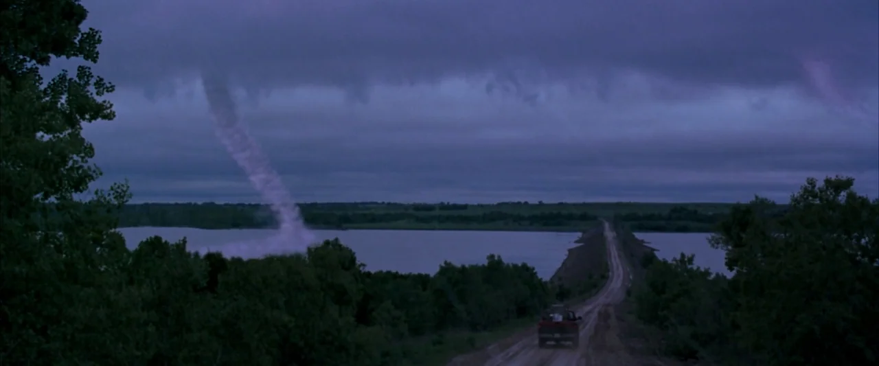 Twister (1996) movie still: establishing — A rural dirt road stretches across a lake under a dark, overcast twilight sky. A single pickup truck…