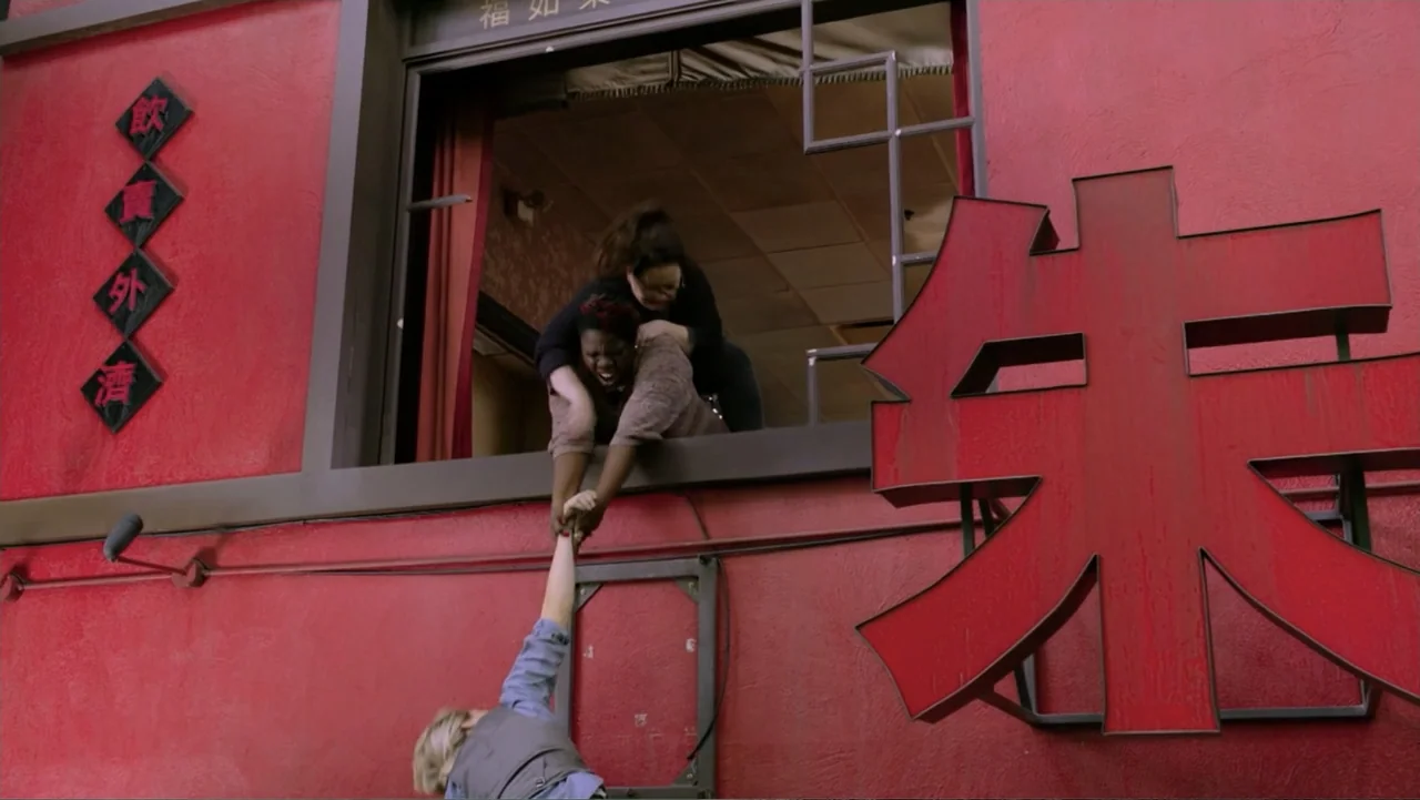 Ghostbusters (2016) movie still: medium — A vibrant red exterior wall of a building features a window and large signs. One person inside the window…