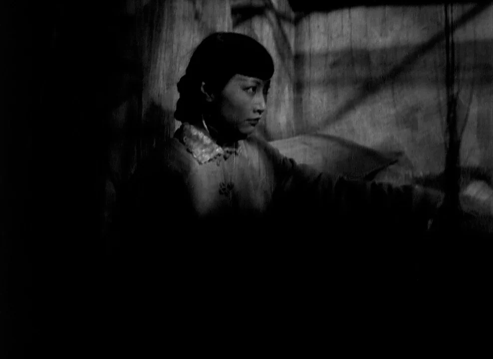 Shanghai Express (1932) movie still: medium — A dark, minimally lit room with wooden wall panels visible in the background. A young woman with a…