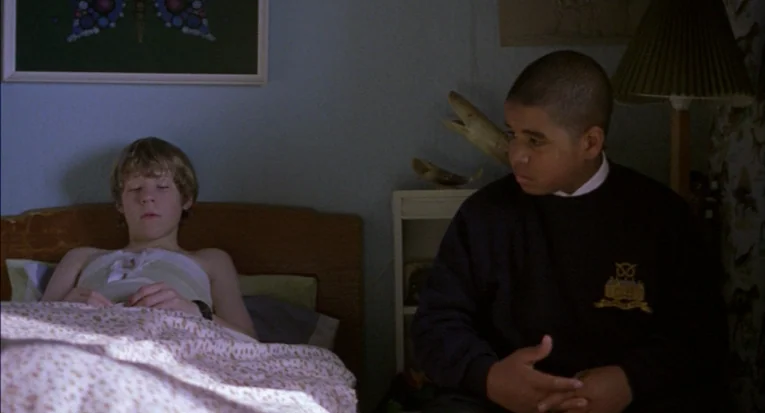 A Room For Romeo Brass (1999) movie still: medium — A bedroom with blue walls and a patterned quilt on the bed. A boy in a dark sweater sits by the…