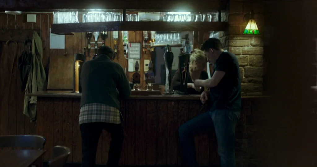 God’s Own Country (2017) movie still: medium — A dimly lit pub features wooden paneling and a long bar with beer taps. Two men converse while…