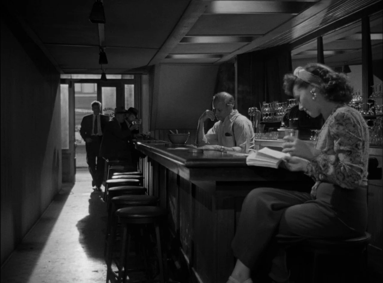 Criss Cross (1949) movie still: medium — A dimly lit bar interior features a long wooden counter and rows of stools. A woman sits reading in the…