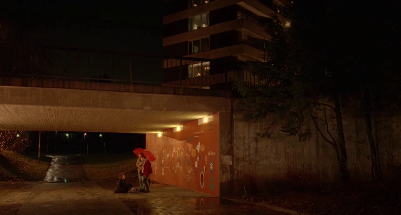 We Are The Best! (2013) movie still: wide — A concrete underpass at night features a brightly painted orange wall covered in graffiti. Three…