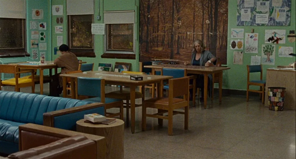 Choke (2008) movie still: wide — A community center room with mint green walls features child-like artwork and wall murals. Two individuals sit at…