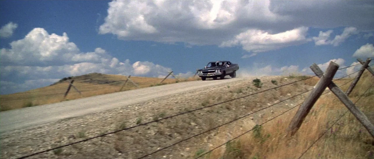 Thunderbolt & Lightfoot (1974) movie still: wide — A dark sedan drives along a dusty, elevated gravel road through arid hills. A barbed wire fence…