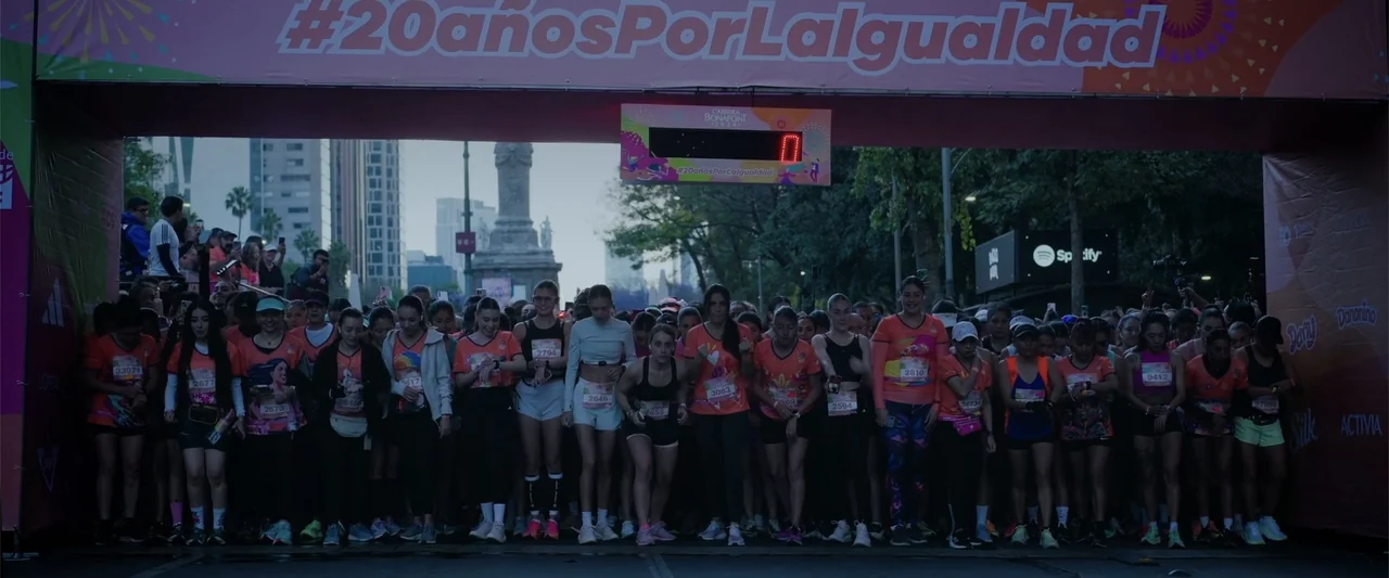 Winner Takes the Cake (2025) movie still: wide — A wide city street is occupied by a large group of runners at the start. A pink archway banner with…