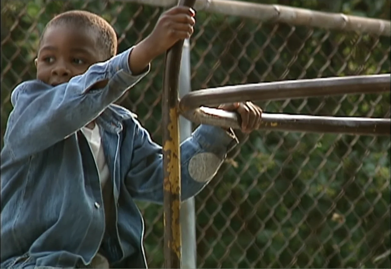 Broken Child (2000) movie still: medium — A young child stands at an outdoor playground near a chain-link fence. The boy grips a weathered metal…