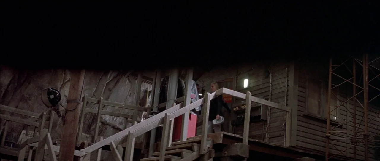 A View to a Kill (1985) movie still: wide — A wooden staircase leads to a small structure built into a rocky hillside. A man climbs the stairs while…
