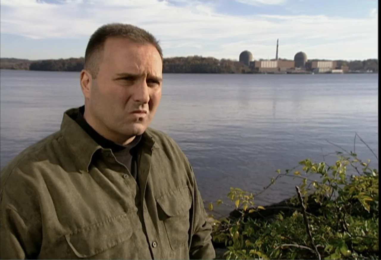 Indian Point: Imagining the Unimaginable (2004) movie still: medium — A man stands on a riverbank with a large nuclear power plant in the distance.…