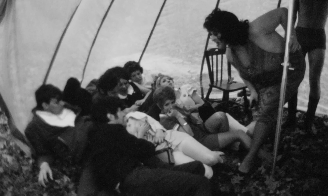 Multiple Maniacs (1970) movie still: medium — People sit on the ground beneath the rounded, light-colored fabric canopy of a tent. A woman stands on…