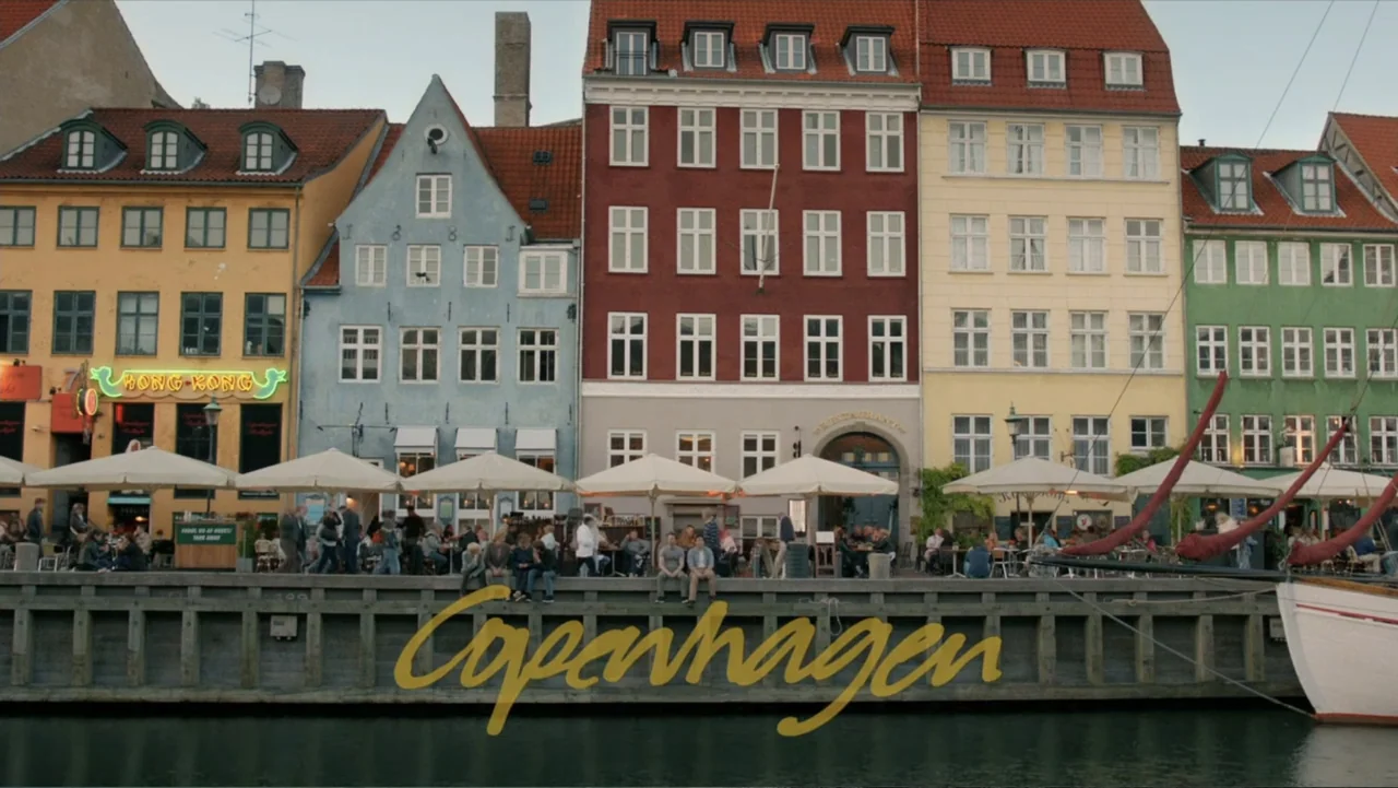 Copenhagen (2002) movie still: wide — A row of colorful historic buildings faces a harbor with a concrete quay. People are seated under umbrellas at…
