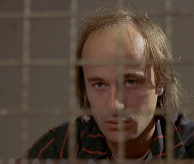 Crime & Punishment (1983) movie still: closeup — A man's face is positioned behind a metal wire grid screen. The background is out of focus and…