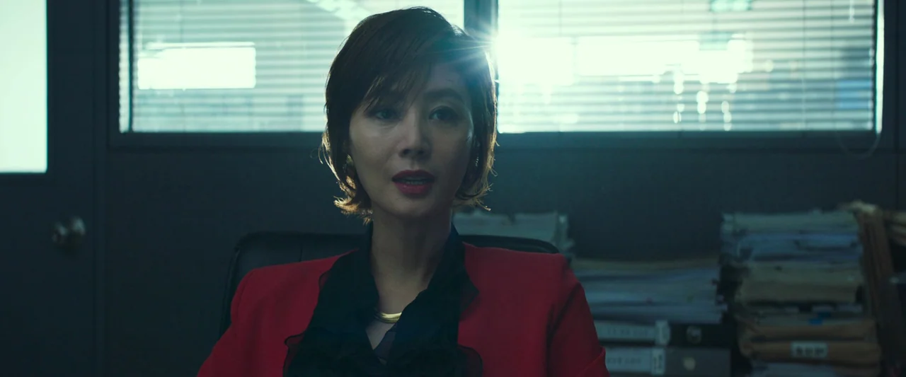 Believer (2018) movie still: medium — An office environment contains piles of folders and binders arranged on a desk surface. A woman in a red…