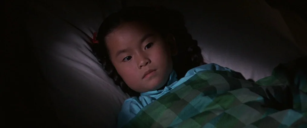 It Happened At The World’s Fair (1963) movie still: closeup — A dark bedroom interior features a young girl lying under a checkered green blanket.…