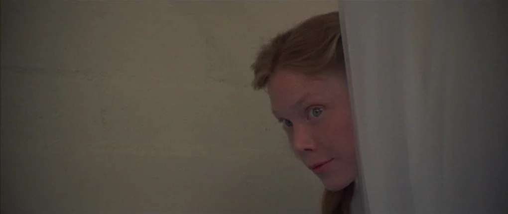 3 Women (1977) movie still: closeup — A dimly lit, plain, beige-walled shower interior. A young woman with reddish-blonde hair peers out from behind…