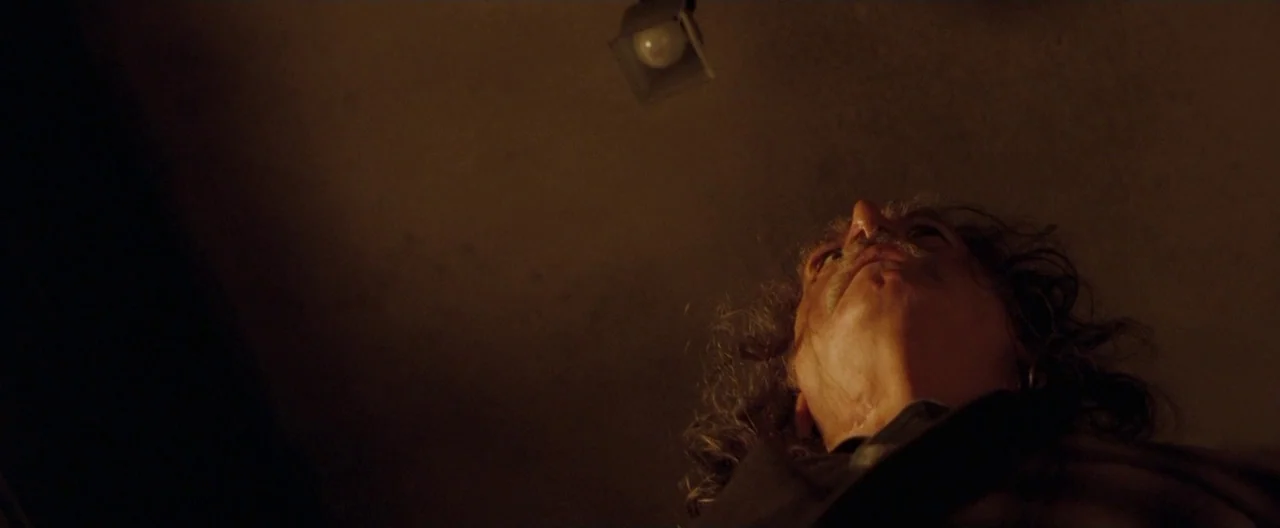 Trick ‘R Treat (2007) movie still: closeup — A dark, minimally lit room with a single light fixture overhead. An older man gazes directly up at the…