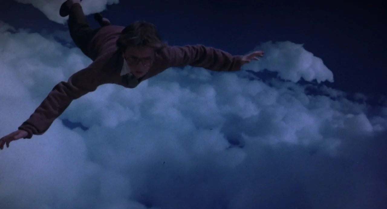 Explorers (1985) movie still: fullbody — A dense layer of clouds spans the frame under a dark blue sky. A young boy in a brown sweater floats…