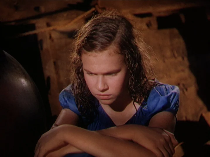 The River (1951) movie still: closeup — A dark, cramped space with wooden planks forming the walls and rafters. A young girl in a blue dress sits…